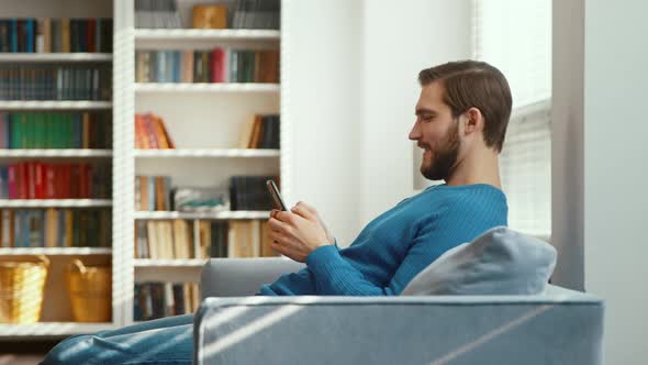 Smiling young man typing on smartphone on sofa