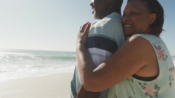 Smiling senior african american couple embracing and looking at sea on sunny beach
