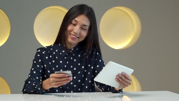 An Asian Woman Using a Bank Card to Pay for Goods Purchased Online Using a Tablet at a Table