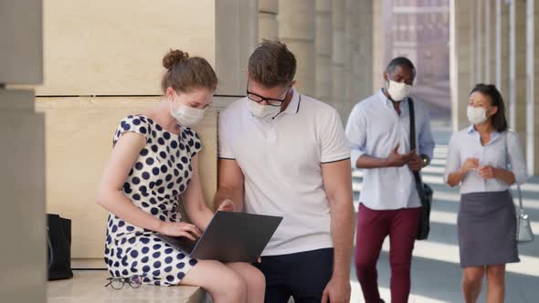 Caucasian Young Colleagues in Medical Mask Sitting Outdoor and Talking While Working on Laptop