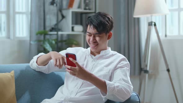 Asian Man Touching Smartphone And Laughing While Lying On Sofa In The Living Room