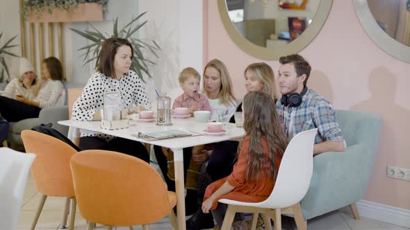 Happy Adult Women, Man and Little Children Are Sitting at Table in Cozy Cafe