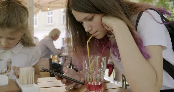 Teenager Girl with Younger Sister Sitting in an Outdoor Cafe Drinking Juice and Using Smartphone