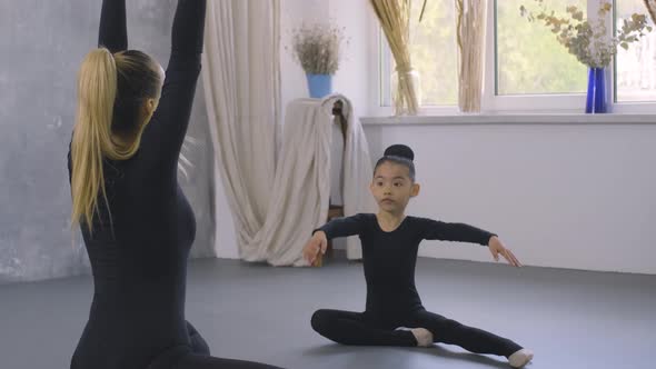 Little Asian Girl in Black Bodysuit Doing Stretching Exercises with the Help of Female Trainer
