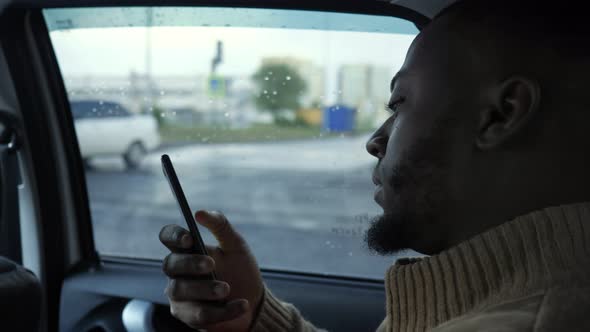 Portrait of Black Man Browsing Smartphone Is Riding a Car in Rainy Day in City.