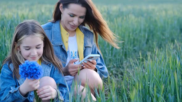 Mom and Daughter Use a Smartphone Sitting in the Tall Green Grass in the Middle of the Field