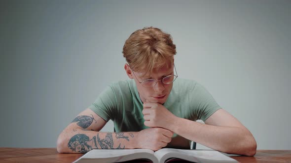 Young Man in Glasses Reading a Textbook and Stressing with His Fist in His Chin