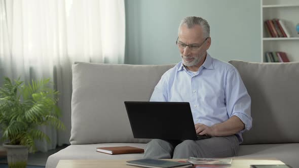 Healthy Senior Male Sitting on Sofa and Viewing Photos on Dating Website, Laptop