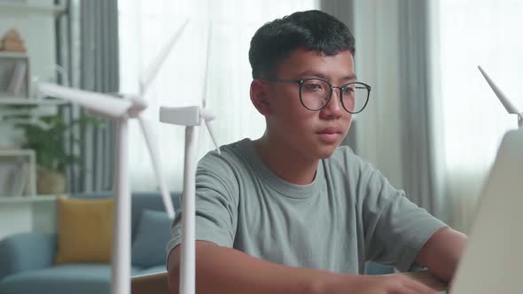 Asian Kid Or Student With Laptop Computer Programming Electric Wind Turbine Model At Home