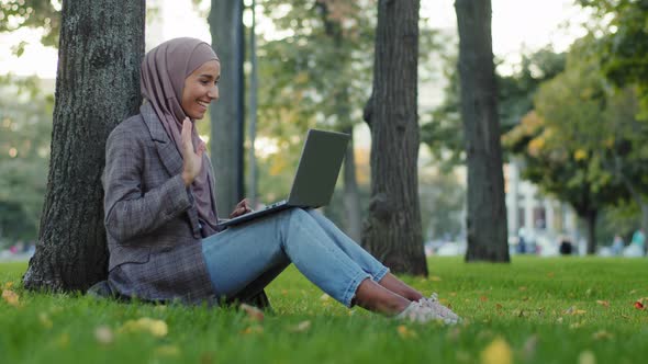 Happy Muslim Islamic Girl Business Woman in Hijab Sitting Near Tree on Green Grass in City Park