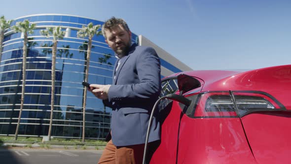 Man in Blue Suit Standing at Charging Electric Car Working on Smartphone