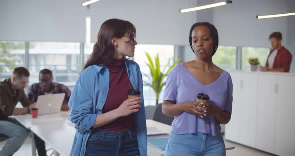 European and African American Women with Cups Smile and Talk in Office Interior