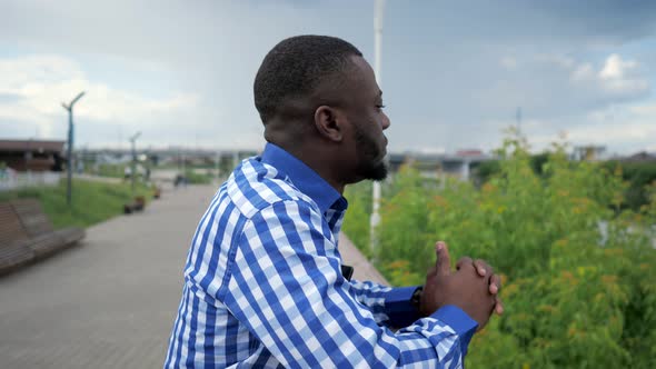 Black Man Stands on City Waterfront Near Fence in Park and Admires City View