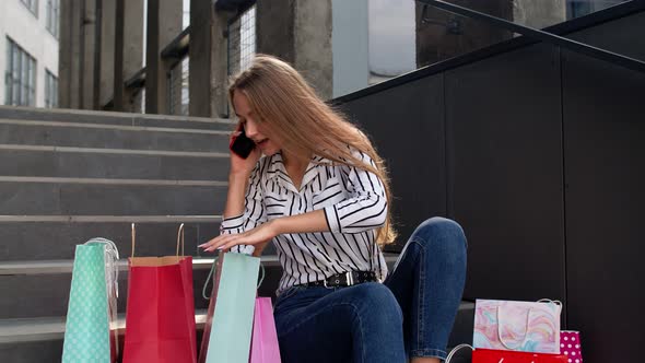 Girl Sitting on Stairs with Bags Talking on Mobile Phone About Sale in Shopping Mall in Black Friday