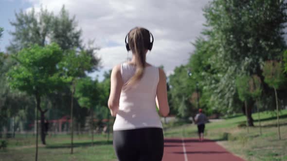 Camera Follows Slim Happy Caucasian Sportswoman in Headphones Jogging on Running Track on Summer