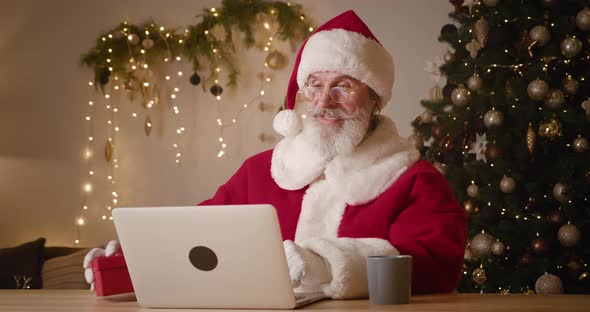 Elderly Bearded Man Dressed As Santa Sitting at Table with Laptop and Talking on Video Call Showing