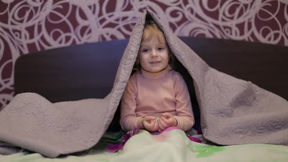 Little Cheerful Girl Hides Under Blanket and Watching TV. Concept of Kids Sleep