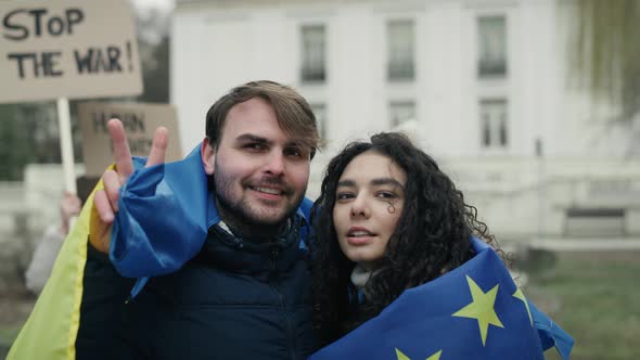 Embraced caucasian man and woman covered with EU and Ukraine flags are showing a peace sign. Shot wi