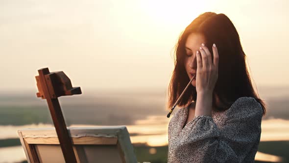 A Young Beautiful Woman Artist Draws a Painting at Sunset Field  Puts Her Hair Above Her Ear