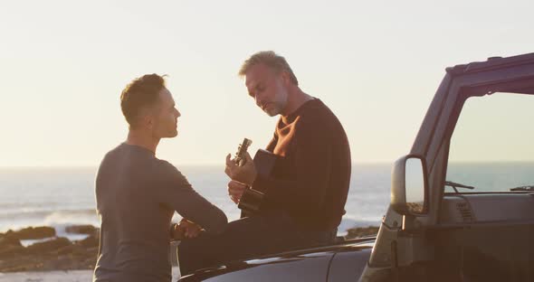 Happy caucasian gay male couple sitting on car playing guitar and talking at the beach