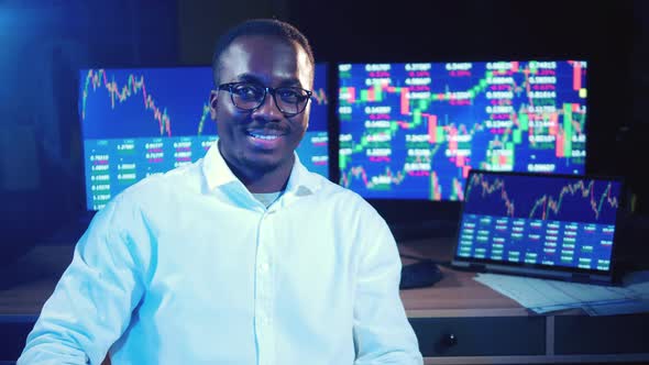 African Broker is Sitting Next to the Monitors with Stock Diagrams