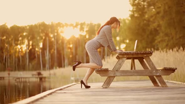 Young Business Woman in Suit Bends Over and Uses Laptop Outdoors During Sunset on Lake
