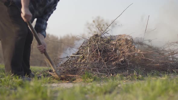 Mature Caucasian Farmer Throwing Dry Leaves and Branches with Shovel Into Fire Outdoors