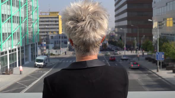 A Middle-aged Woman Stands on a Bridge and Looks Down at the Busy Street and Cityscape - Closeup