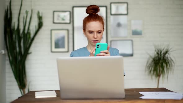 Portrait of Confused Redhead Young Woman Typing on Mobile Phone Sitting at Desk.