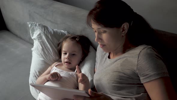 Mom and Little Daughter Are Lying in Bed with a Wake Cartoon on a Tablet. Mother and Baby Make Video