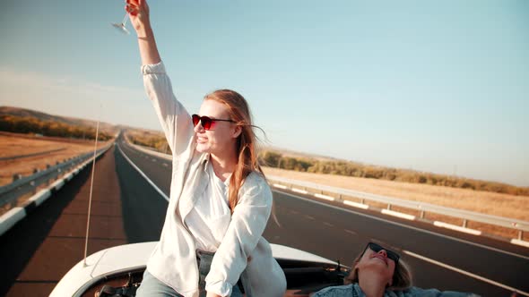 Two female travelers in back seat of car with an open roof raise glasses of alcohol at hands, drivin