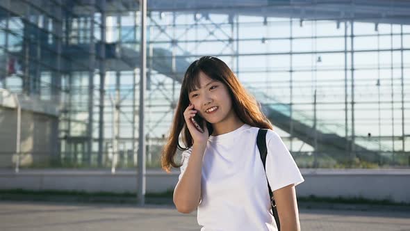 Smiling Young Asian Girl with Dark Long Hair Talking on Mobile Near the Airport in Day Time