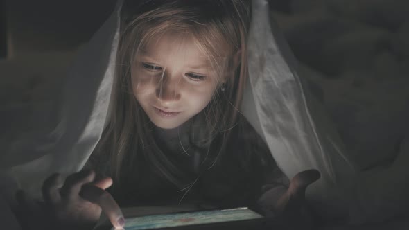 Teenage Girl in Bed Playing a Tablet in Social Internet in the Dark Light. Close Up of Little Girl