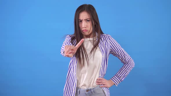 Young Sullen Asian Woman Stands on a Blue Background and Points Her Finger in Denial