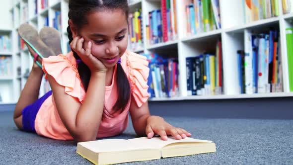 Schoolgirl lying on floor and reading a book in library at school