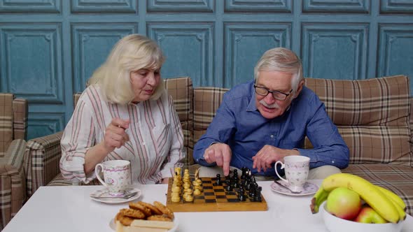 Smiling Senior Couple Grandfather Grandmother Resting on Sofa Drinking Coffee Playing Chess at Home