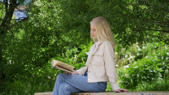 Carefree Young Woman Reading a Fascinating Novel While Sitting on a Park Bench