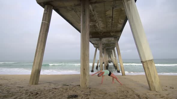 A young attractive woman doing yoga on the beach under a pier