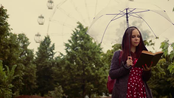 Young Beautiful Woman with Transparent Umbrella Reading Book Standing in City Park in Rain