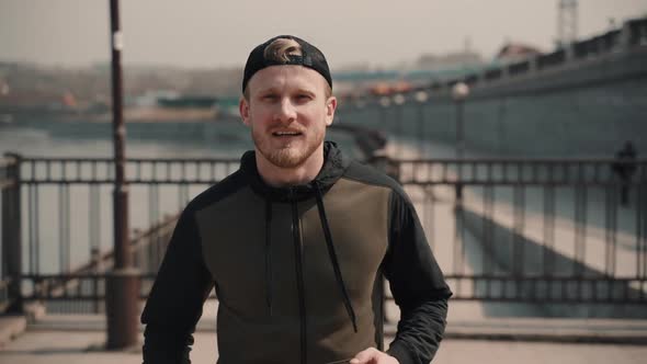 Front View of Young Bearded Man Is Running on City Bridge in Spring Afternoon.