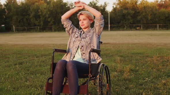 Disabled Woman is Sitting in Wheelchair on Lawn with Her Hands Raised Above Head She Lowers Her