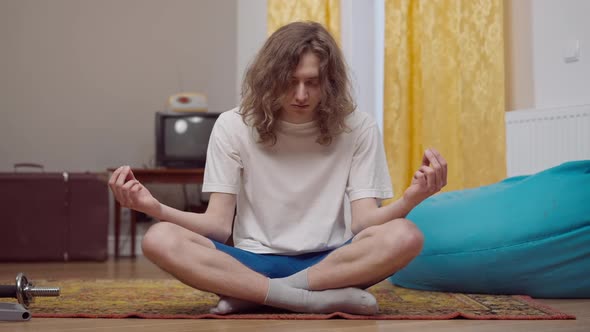Portrait of Calm Young Retro Man with Long Curly Hair Falling Asleep Sitting in Lotus Pose Indoors