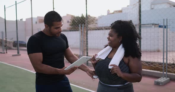 Curvy african woman talking with her personal trainer while checking tech clipboard outdoor