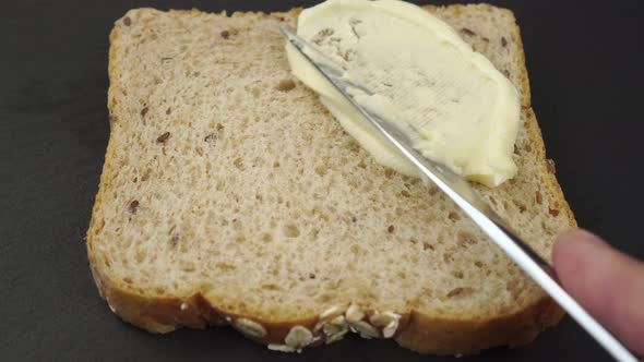 Butter is spread with a knife over a slice of cereal bread on a gray stone surface. 