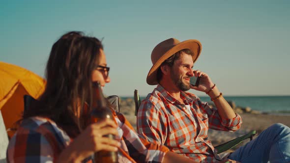 Happy Carefree Businessman with Wife or Girlfriend Relaxing with Beer in Camp Chairs Next to Tent on