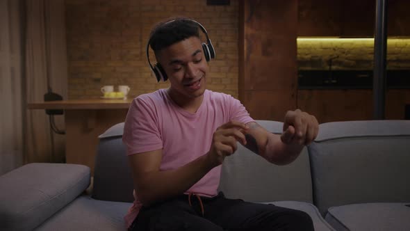 20s African American Man Enjoys Listening the Music in Wireless Headphones Sitting on Sofa at Home