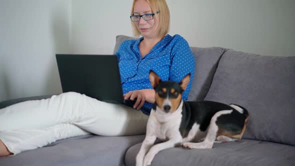 A Woman Working on a Laptop From Home While Sitting on a Couch with Her Cute Dog