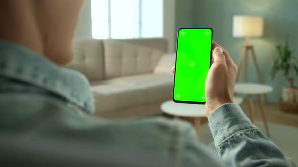 Back View of Young Man at Home Sitting on a Chair Using With Green Mock-up Screen Smartphone. Boy Is