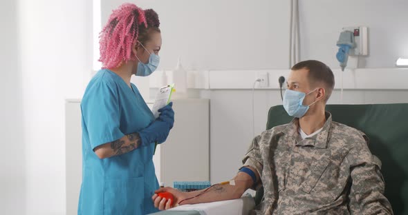 Nurse in Safety Mask Talking to Soldier Sitting on Chair Receiving Blood Transfusion at Hospital