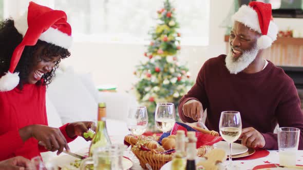 African american family in santa hats talking and smiling while sitting on dining table having lunch